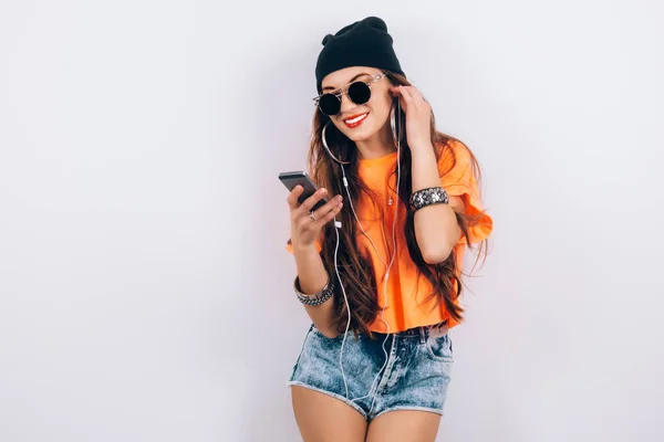 Junge Hipster schöne Frau mit Sonnenbrille, schwarzem Hut und orangefarbenem T-Shirt, die Musik über Kopfhörer in der Nähe der weißen Wand hört, mit einem Handy in der Hand — Stockfoto