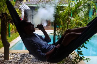Bir hamakta dinlenen ve elektronik sigaradan buhar salan adam