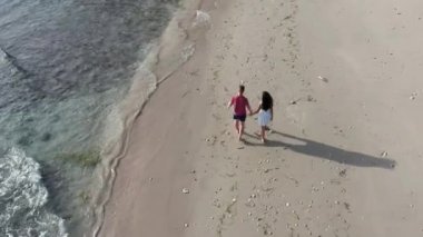 Gün batımında Tropik Plajboyunca Yürüyen Erkek ve Kadın, tatilde sahilde eğlenen genç çiftin havadan çekilmiş fotoğrafını takip ediyor. Gün batımında sahilde boş sahilde yürüyen çift.
