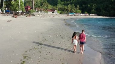Gün batımında Tropik Plajboyunca Yürüyen Erkek ve Kadın, tatilde sahilde eğlenen genç çiftin havadan çekilmiş fotoğrafını takip ediyor. Gün batımında sahilde boş sahilde yürüyen çift.
