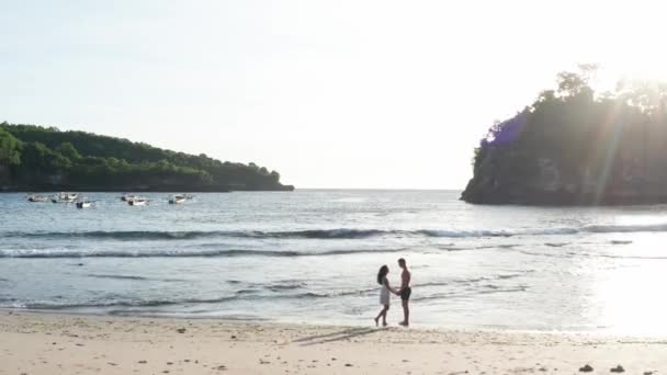 男人和女人在日落时沿着热带海滩散步，跟踪年轻夫妇在度假的航拍，在海滩上玩得开心。情侣在日落时分在海滩的空沙滩上散步. — 图库视频影像