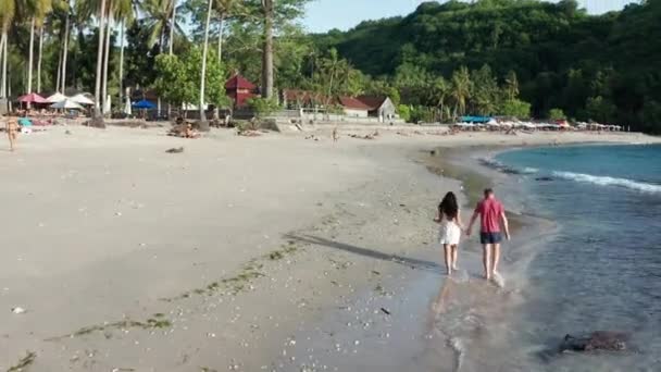 Hombre y mujer caminando por la playa tropical al atardecer, rastreando la toma aérea de una joven pareja de vacaciones divirtiéndose en la playa. Pareja caminando en la playa vacía al atardecer junto a la playa . — Vídeo de stock