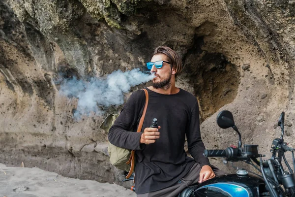 Gün batımı plajda açık havada sigara elektronik sigarette motosiklet üzerinde sakallı ve güneş gözlüğü koltuk ile Adam. Genç hipster erkek deniz kenarında vaping.