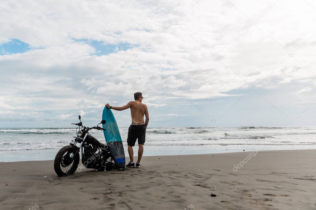 male person looking for inspiration while standing near sportive motorbike. holding surfboard in hands. Young man surfer enjoying recreation near to ocean.