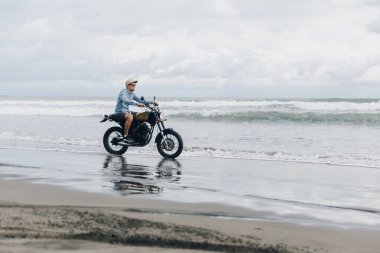 Sahilde motosiklete binen şapkalı adam. Bali plajda gün batımında Moto çapraz dirtbiker. Genç hipster erkek özgürlük ve aktif yaşam tarzı zevk, bir bisikletçiler tur eğlenmek.