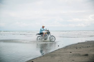 Sahilde motosiklete binen şapkalı adam. Bali plajda gün batımında Moto çapraz dirtbiker. Genç hipster erkek özgürlük ve aktif yaşam tarzı zevk, bir bisikletçiler tur eğlenmek.