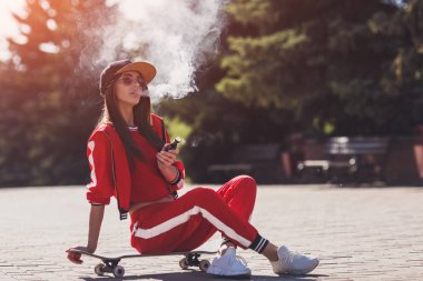 Vaping girl. Young woman sitting on skateboard and vape e-cig. Pretty young female in black hat, red clothing vape ecig, vaping device at the sunset. Toned image. Hip-hop style.