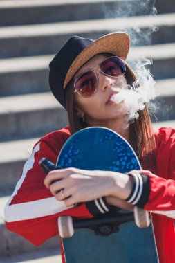 Vaping girl. Young woman with skateboard vape e-cig. Pretty young female in black hat, red clothing vape ecig, vaping device at the sunset. Toned image. Hip-hop style.