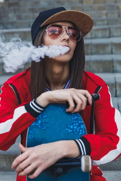 Vaping lány. Fiatal nő gördeszka VAPE e-cigi. Csinos fiatal nő a fekete kalap, piros ruha VAPE ecig, gőzös eszköz a naplementében. Tónusú kép. Hip-hop stílus. — Stock Fotó