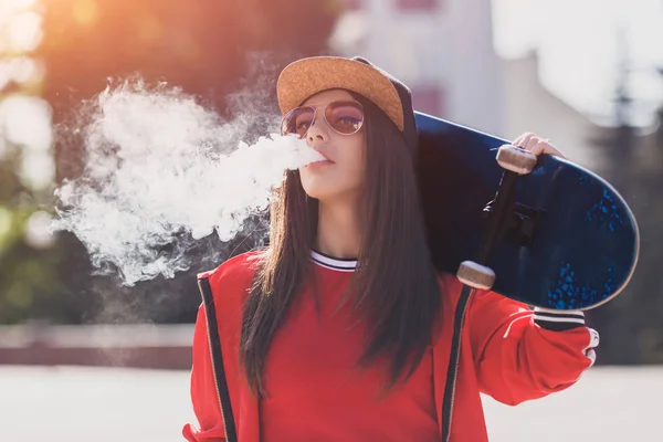 Vaping lány. Fiatal nő gördeszka VAPE e-cigi. Csinos fiatal nő a fekete kalap, piros ruha VAPE ecig, gőzös eszköz a naplementében. Tónusú kép. Hip-hop stílus. — Stock Fotó