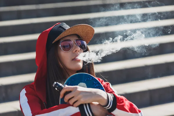 Vaping girl. Jeune femme avec skateboard vape e-cig. Jolie jeune femelle en chapeau noir, vêtements rouges vape ecig, dispositif de vapotage au coucher du soleil. Image tonique. Style hip-hop . — Photo