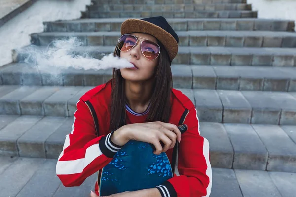 Выпендривающаяся девушка. Молодая женщина со скейтбордисткой электронной сигаретой. Красивая молодая женщина в черной шляпе, красная одежда, рецепт испарения, устройство для испарения на закате. Тонированное изображение. Хип-хоп стиль . — стоковое фото