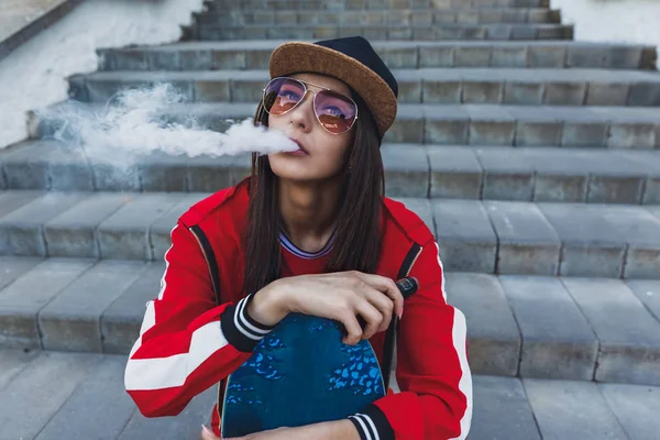 Gadis Uap. Wanita muda dengan skateboard vape e-cig. Wanita cantik bertopi hitam, pakaian merah, ecig vape, alat pengap saat matahari terbenam. Citra berdada. Gaya hip-hop . — Stok Foto