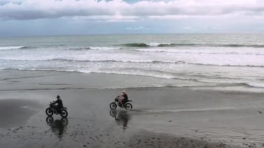 Okyanus ayakın siyah kum plajda modern özel motosiklet kafe yarışçı sürme iki yakışıklı hipster erkek. Macera seyahat konsepti. Okyanus dalgalarıyla sörf noktası. Havadan ateş. 4k