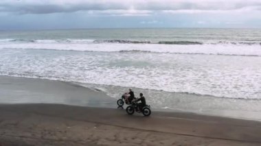 Okyanus ayakın siyah kum plajda modern özel motosiklet kafe yarışçı sürme iki yakışıklı hipster erkek. Macera seyahat konsepti. Okyanus dalgalarıyla sörf noktası. Havadan ateş. 4k