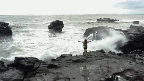Kolları tamamen açık özgürlüğü olan kadın kayanın üzerinde duruyor. Havadan ateş. Kayanın üzerinde duran genç beyaz kadın fırtınalı okyanus denizine bakıyor dalgalarla deniz kıyısına çarpar. 4k — Stok video