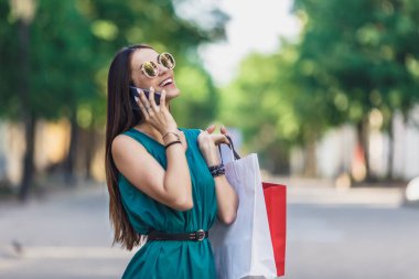 Diş ile gülümseyen ve telefonda konuşan alışveriş çantaları ile genç büyüleyici koyu saçlı beyaz kadın yakın. Pozitif duygular ve alışveriş günü konsepti.