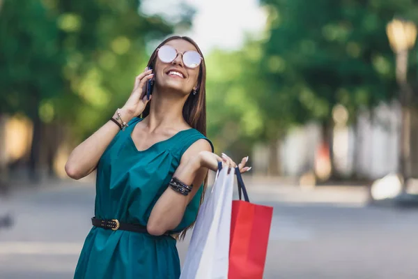 Close up of young charming dark-haired caucasian female with shopping bags smiling with teeth and talking on phone. Positive emotions and Shopping day concept.