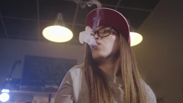 Uma jovem bonita de boné vermelho fuma um cigarro eletrônico na loja de vape. Fecha a porta. Movimento lento . — Vídeo de Stock