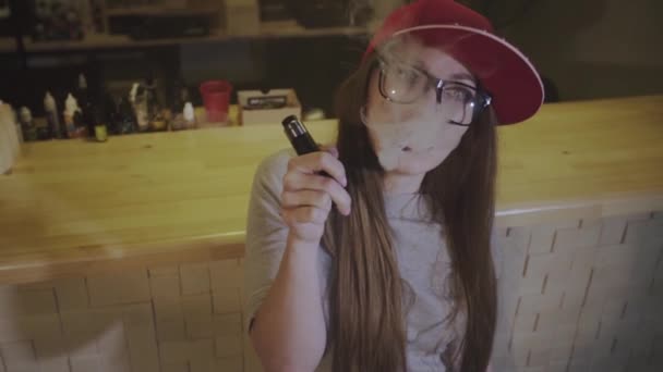 Uma jovem bonita de boné vermelho fuma um cigarro eletrônico na loja de vape. Fecha a porta. Movimento lento . — Vídeo de Stock