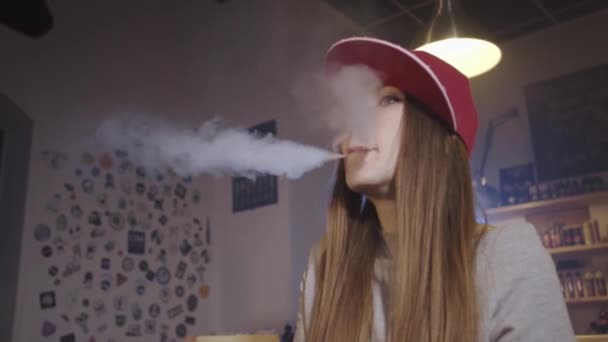 Νεαρή όμορφη γυναίκα με κόκκινο καπέλο καπνίζει ηλεκτρονικό τσιγάρο στο μαγαζί με τα βιντεάκια. Κοντινό πλάνο. Αργή κίνηση. — Αρχείο Βίντεο