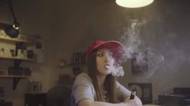 Una joven guapa con gorra roja fuma un cigarrillo electrónico en la tienda de vapores. Primer plano. Movimiento lento . — Vídeos de Stock