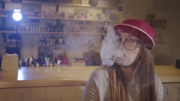 Uma jovem bonita de boné vermelho fuma um cigarro eletrônico na loja de vape. Fecha a porta. Movimento lento . — Vídeo de Stock