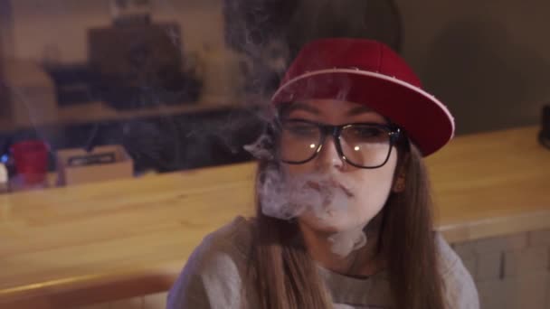 Uma jovem bonita de boné vermelho fuma um cigarro eletrônico na loja de vape. Fecha a porta. Movimento lento . — Vídeo de Stock