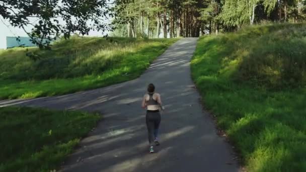 शहर पार्क में दौड़ने वाली युवा आकर्षक लड़की। लकड़ी के हवाई बैक व्यू में दौड़ने वाली महिला। 4K, ट्रैकिंग शॉट . — स्टॉक वीडियो