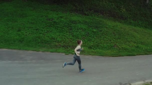 Giovane ragazza attraente in esecuzione nel parco della città. Donna che corre in legno aerea vista laterale. 4K, inseguimento tiro . — Video Stock