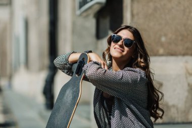 Yakın Portre Güzel genç kadın yerde oturan ve şehir sokak güneşli havabir longboard tutarak. Genç hipster kız kaykay ile poz.