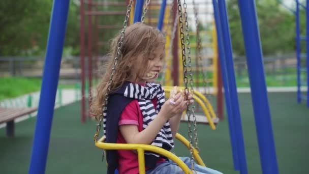 Маленькая девочка сидит на качелях и ест сэндвич на детской площадке. Летний ветреный день. Закрытие и карантин закончились. . — стоковое видео
