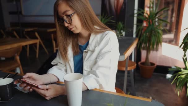 迷人的女人用智能手机戴眼镜，在咖啡店喝咖啡的镜头。女性在社交媒体上发短信和分享信息，享受移动技术，在咖啡店休息. — 图库视频影像