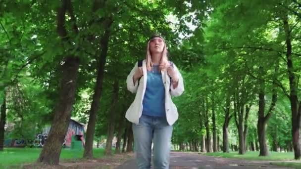 Vista frontal de una hermosa mujer en gorra caminando por el parque con mochila . — Vídeo de stock