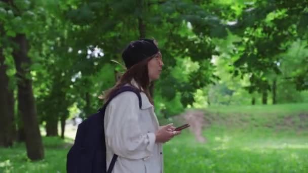 Vue latérale d'une belle femme en casquette marchant dans le parc avec sac à dos et téléphone portable . — Video