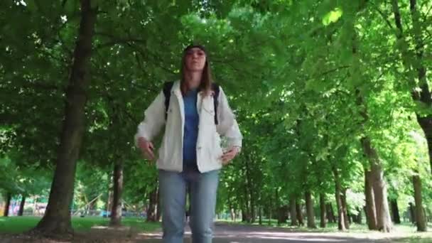 Vista frontal de una hermosa mujer en gorra caminando por el parque con mochila . — Vídeos de Stock