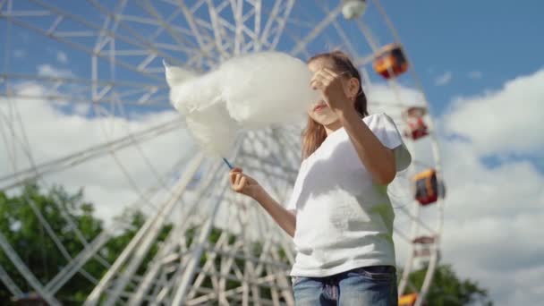 Ritratto di bambino con zucchero filato dolce. Una bambina sullo sfondo della ruota panoramica sta mangiando caramelle-filo interdentale giornata estiva soleggiata . — Video Stock