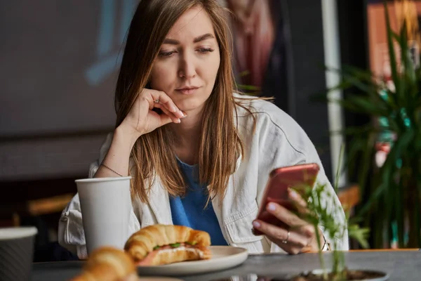 Front view of attractive woman wearing glasses using smartphone, drinking coffee in cafe. Female texting and sharing messages on social media, enjoying mobile technology, relaxing in coffee shop. — Stock Photo, Image