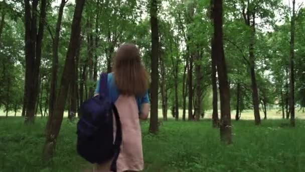 Krásná žena se usmívá po pěšině obklopené listím a stromy. Zadní pohled na fenku s batohem kráčející v letním lese. — Stock video