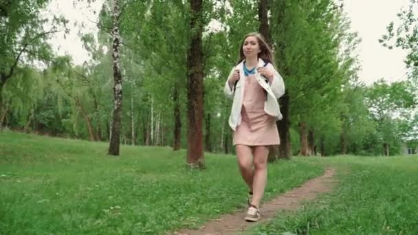 Gyönyörű nő mosolyog sétálni az úton körül lombozat és fák. Elülső kilátás a női hátizsák séta a nyári parkban. — Stock videók