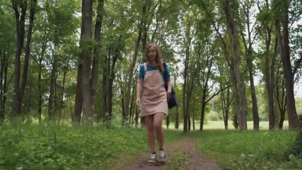 Hermosa mujer sonríe caminando por el camino rodeado de follaje y árboles. Vista frontal de la hembra con mochila caminando en el bosque de verano . — Vídeos de Stock