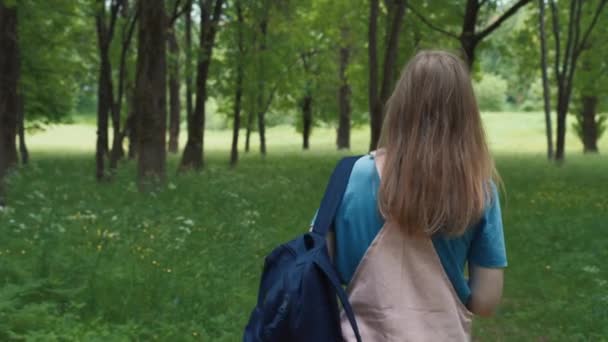 Krásná žena se usmívá po pěšině obklopené listím a stromy. Zadní pohled na fenku s batohem kráčející v letním lese. — Stock video