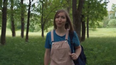 Güzel bir kadın ağaçlar ve yapraklarla çevrili patikada yürürken gülümsüyor. Sırt çantalı bir kadın yaz ormanında yürüyor..