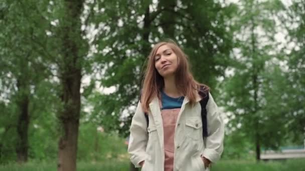 Belle femme sourit en marchant le long du chemin entouré de feuillage et d'arbres. Vue de face de la femelle avec sac à dos marchant au parc d'été. — Video