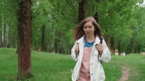 美丽的女人微笑着沿着被树叶和树木环绕的小径走着。带着背包在夏季公园散步的女性的前景. — 图库视频影像