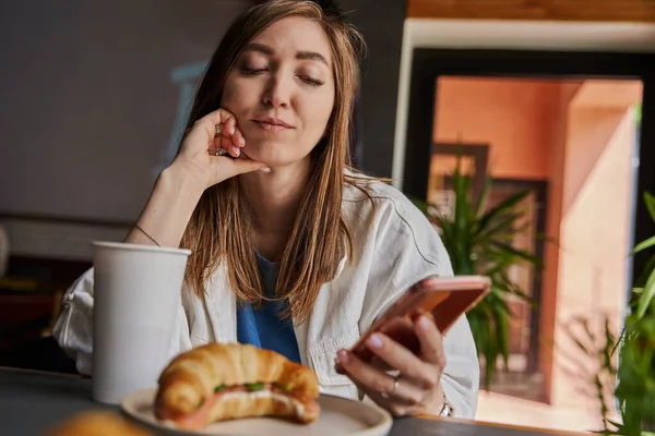 Front view of attractive woman wearing glasses using smartphone, drinking coffee in cafe. Female texting and sharing messages on social media, enjoying mobile technology, relaxing in coffee shop. — Stock Photo, Image