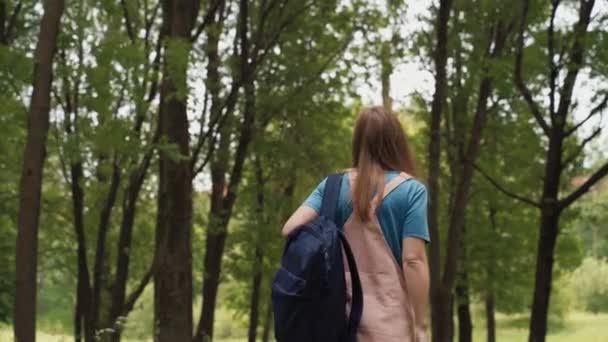 Piękna kobieta uśmiecha się idąc ścieżką otoczona liśćmi i drzewami. Widok z tyłu samicy z plecakiem spacerującym po letnim lesie. — Wideo stockowe