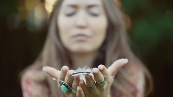 Krásná mladá žena v bohostyle oblečení pózovat v paprscích večerního slunce, západ slunce. Boho styl módy, ženské foukání peří v parku venku. Zavřít. Selektivní zaměření. — Stock video