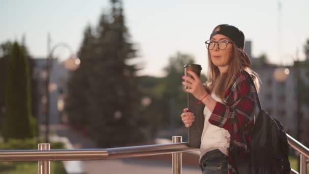 Feliz joven estudiante vestida con ropa casual con taza de café y mochila a sus espaldas caminando por la ciudad. Estudiante sosteniendo laptop y bebiendo café. Hora del atardecer . — Vídeos de Stock