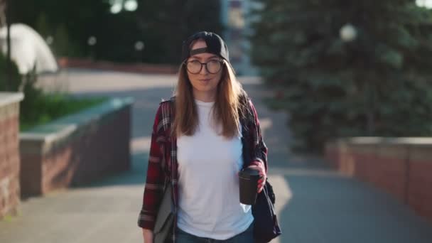 Gledelig, ung kvinnelig student kledd i løse klær med kopp kaffe og ryggsekk bak ryggen går rundt i byen. Kvinnestudent som holder laptop og drikker kaffe. Sommersolnedgang. – stockvideo